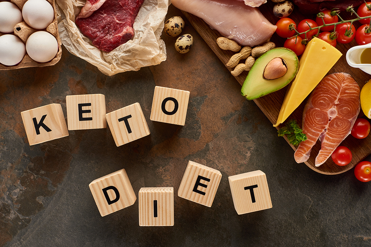 Top view of raw meat and fish near eggs, cheese, tomatoes, avocado and peanuts on marble surface with wooden cubes keto diet inscription
